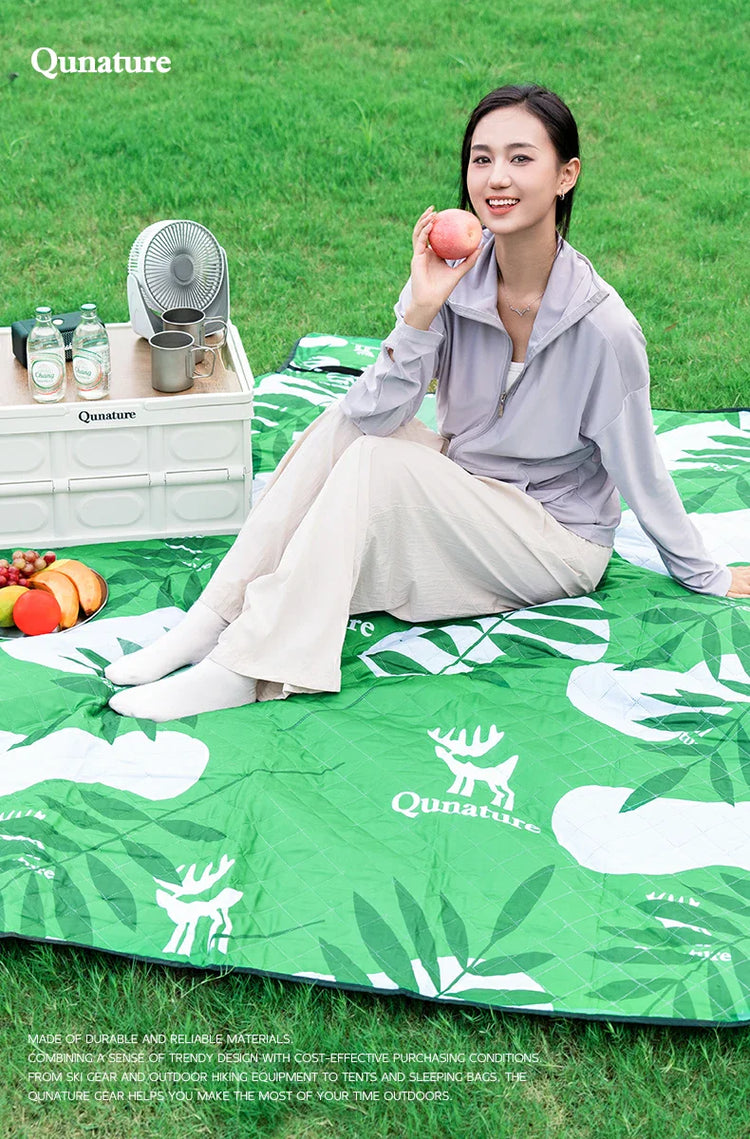 A girl is sitting on a mat on the grass outdoors and smiling, holding an apple in her hand