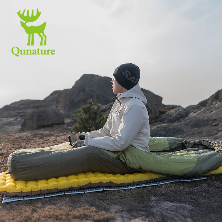 A boy wearing a hat sitting on a sleeping mat in a sleeping bag outdoors looking into the distance