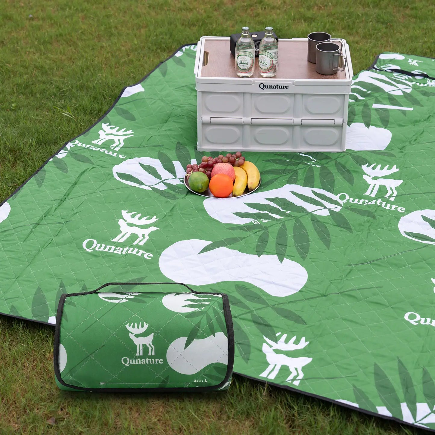 Green picnic mat with white deer and leaf patterns, accompanied by a cooler and food items on grass.