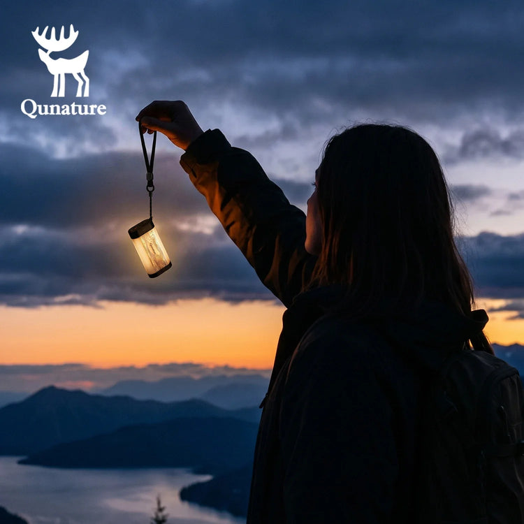 A person in a dark jacket holds up a glowing lantern against a dramatic mountain sunset sky. The scene includes a cloudy sky, distant mountains, and water, with the Qunature logo and deer icon visible in the upper left.