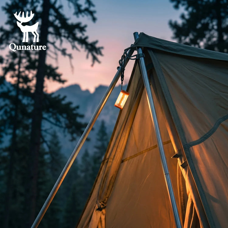 A Qunature brand tent pitched outdoors in a forest at dusk, with a warm lantern hanging from the peak and mountains visible in the background.