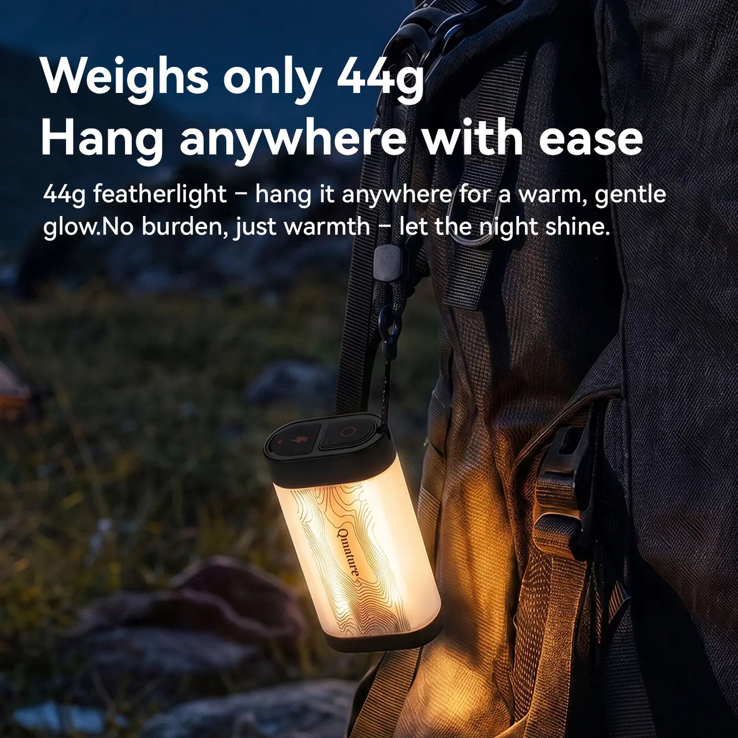 A small cylindrical featherlight lantern hangs from a black backpack outdoors at dusk. The lantern is illuminated with a warm white glow, and white marketing text on the left reads 'Weighs only 44g Hang anywhere with ease' and '44g featherlight – hang it anywhere for a warm, gentle glow.