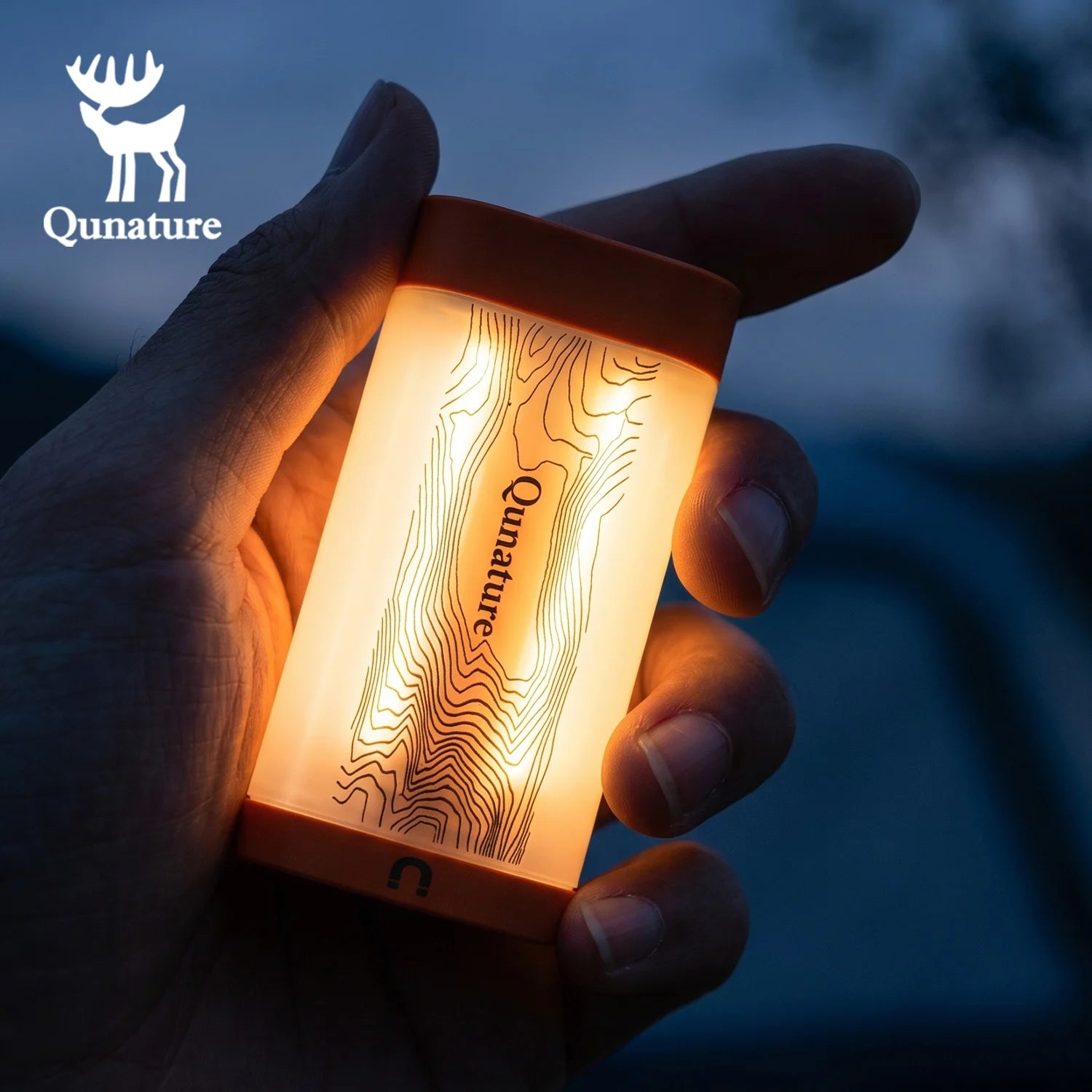 A hand holding a small rectangular Qunature LED lantern that glows with a warm orange light, showing a topographic line pattern on its front. The Qunature brand logo is visible in the top left corner.