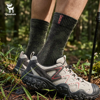 A close-up of a person's lower legs wearing dark charcoal gray ribbed socks with red 'Mountain' branding and off-white hiking shoes with red accents, standing in a forest setting.