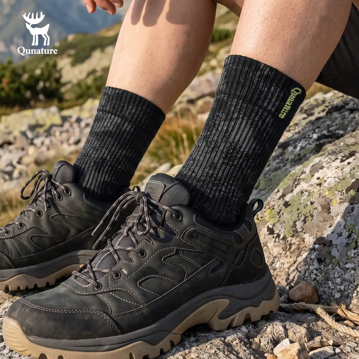 A person sitting outdoors on rocky ground wearing dark hiking shoes and ribbed black crew socks with a green Qunature logo. The Qunature brand logo is also shown as an overlay in the top left corner.
