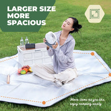 Woman sitting on a large picnic blanket with a fan, surrounded by a picnic setup on a grassy field.