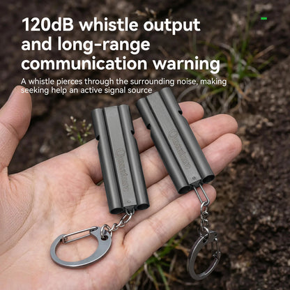 Two dark gray cylindrical whistle keychains held in a person's hand, with outdoor soil and grass in the background and overlaid text about 120dB whistle output and long-range communication warning.