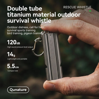 A person's hand holding a compact metallic outdoor survival whistle with a key ring, shown with promotional text on a blurred green background. The text highlights features such as double tube titanium material, 120 dB sound, 14 g weight, and 5.