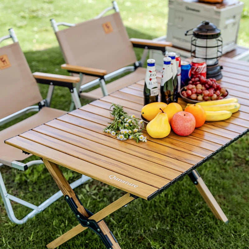 Wooden camping table with fruits, drinks, and chairs on a grassy area
