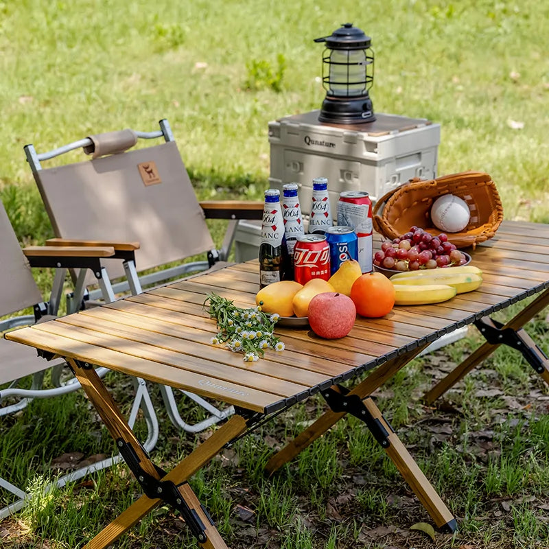 Camping scene with a wooden table, chairs, cooler, and snacks outdoors.