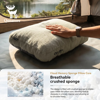 A Qunature cloud memory pillow in a textured beige linen-like fabric, shown with a hand resting on it to demonstrate its softness. The background shows a cozy camping setting with a tent and a lake, and there is a text overlay at the bottom explaining the breathable crushed sponge core.