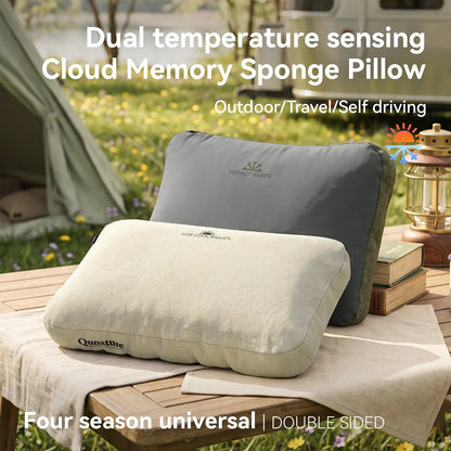 Two Qunature memory foam pillows displayed outdoors on a wooden table, with one in light beige in front and one in dark gray behind. The background shows a blurred camping scene with a tent, grass, and flowers.