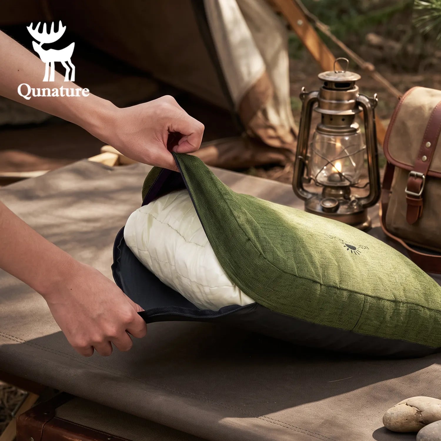 A person's hands are shown opening a green quilted camping pillow case to reveal a white quilted camping pillow, set on a camping cot with a vintage lantern and tent in the background. The Qunature brand logo is visible in the top left corner.