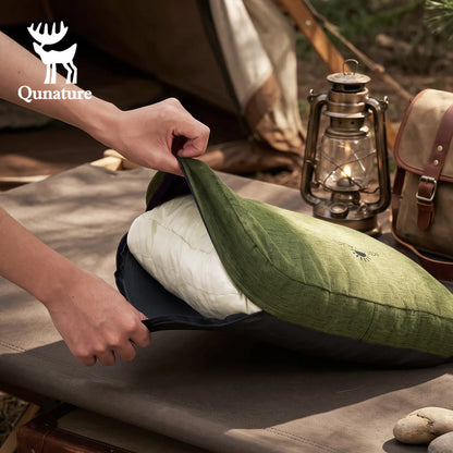 A person's hands are shown opening a green quilted camping pillow case to reveal a white quilted camping pillow, set on a camping cot with a vintage lantern and tent in the background. The Qunature brand logo is visible in the top left corner.