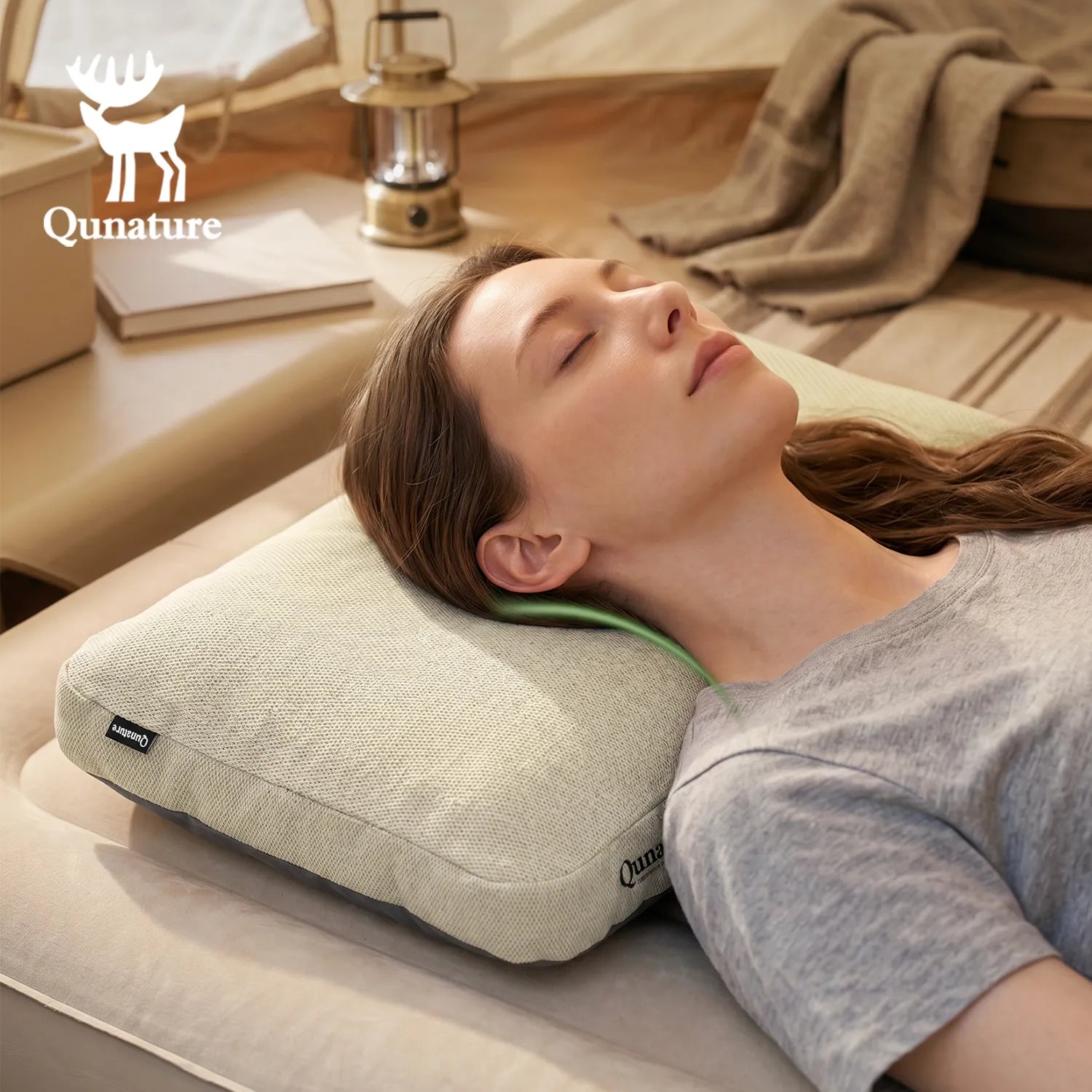 A woman lies on her side on a Qunature pillow in a softly lit bedroom setting. The pillow is light beige with a visible green inner support edge, and the Qunature brand name and deer logo appear in the upper left corner.