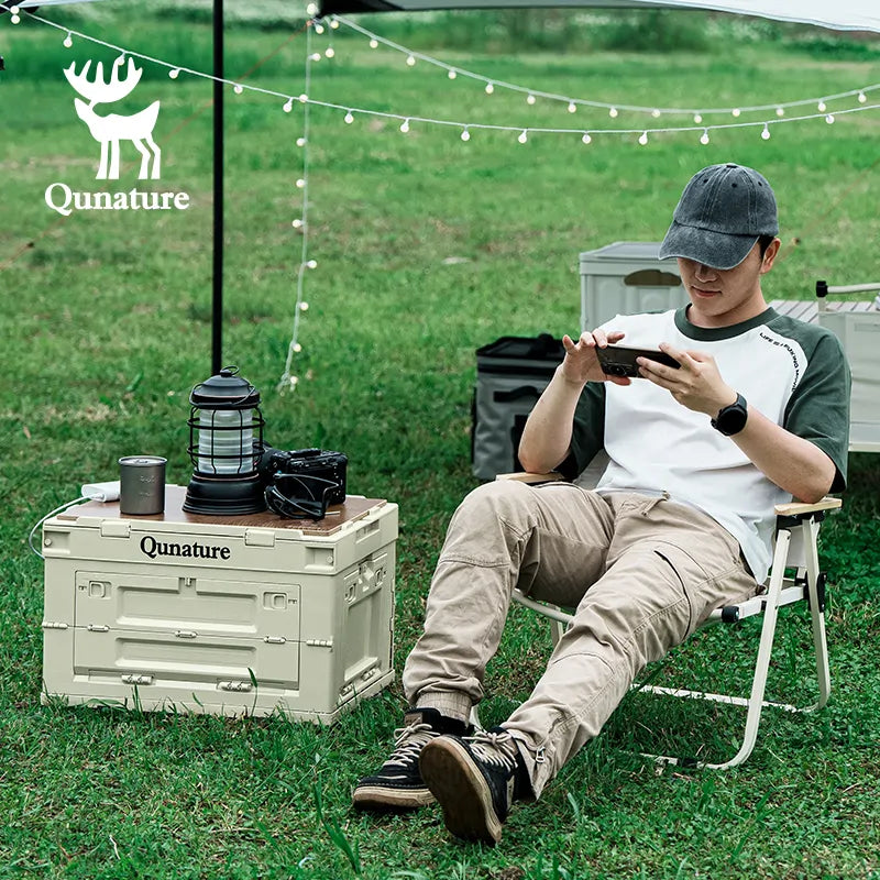 Person sitting outdoors with camping gear, including a Qunature branded crate and lantern.