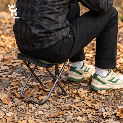 A person sits on a small black folding camp stool outdoors on a ground covered with dry brown leaves. The person wears a dark plaid jacket, black pants, white socks, and green-and-white sneakers.