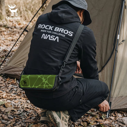 A man in a black outdoor jacket with 'NASA' and 'ROCK BROS' printed on the back, crouching in front of a tent while wearing a green camouflage-patterned sling bag across his chest. The Qu nature logo is visible in the top left corner.