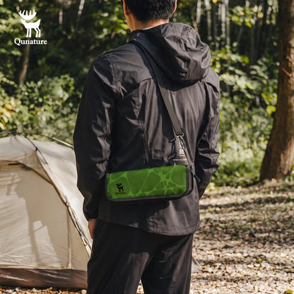 A man wearing a black outdoor jacket stands in a forest campsite, viewed from behind, with a green and black Qunature crossbody bag worn across his chest. A beige tent is visible in the background on a gravel path.