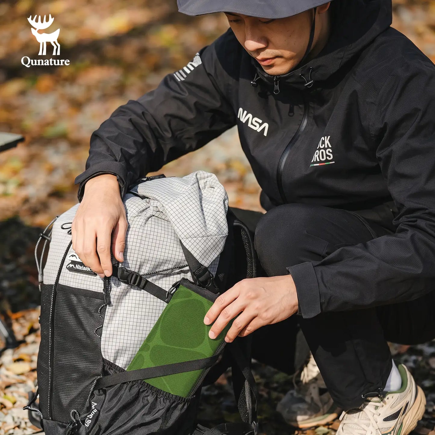 A man in outdoor gear kneels on a forest floor, demonstrating a green and gray backpack. The backpack features a gray and white checkered pattern on the top, a black lower section, and a green side pocket.