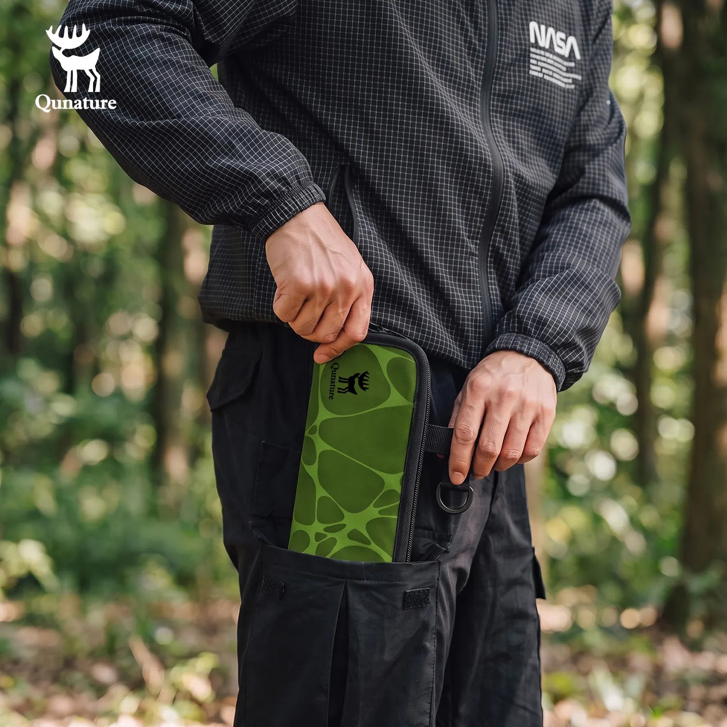 A person in a forest setting wearing a black patterned jacket and black cargo pants, demonstrating the Qunature brand by pulling a green and black patterned pouch from their pocket.