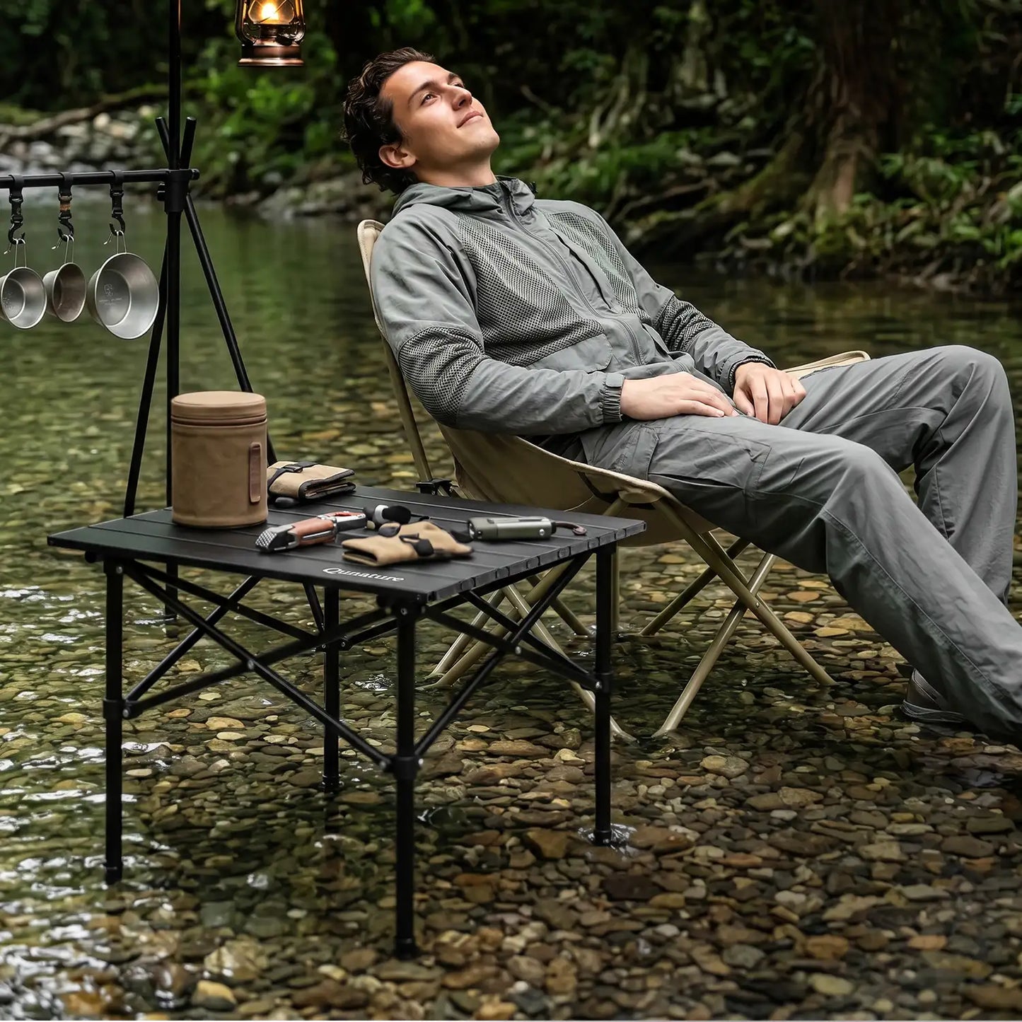 Man sitting by a river with camping gear, including a table and pots, in a natural setting.