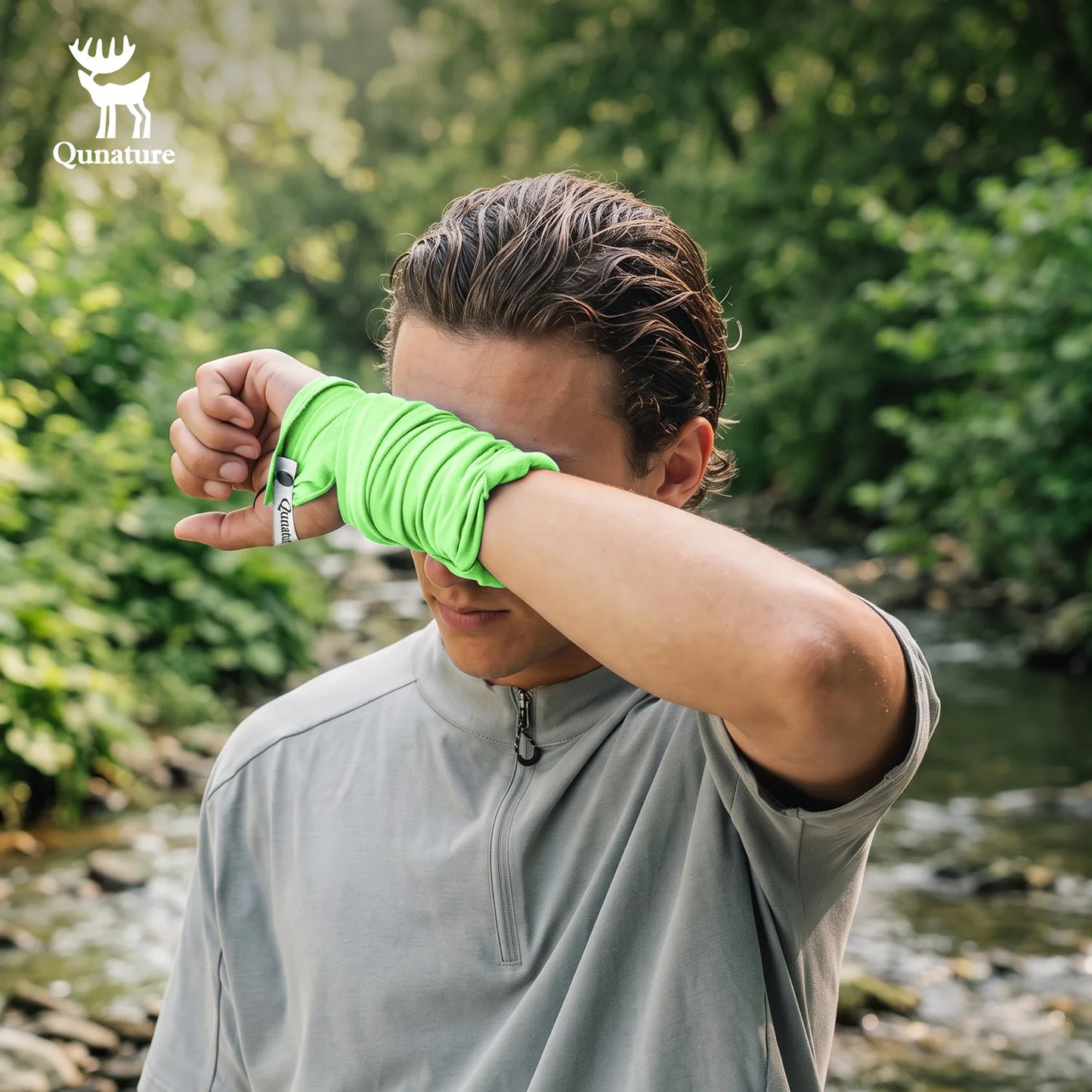 A man outdoors by a stream wearing a bright neon green wrap-around wrist/forearm sleeve over a light gray athletic quarter-zip top. The Qunature brand logo is visible in the top-left corner.