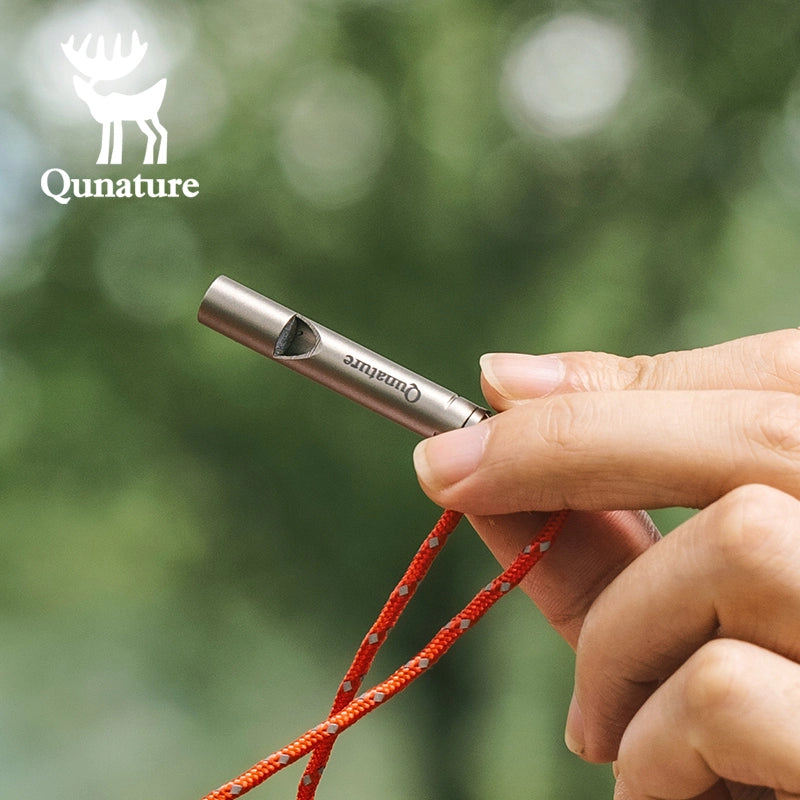 Hand holding a metal whistle with a red cord against a blurred green background, featuring the Qunature brand.