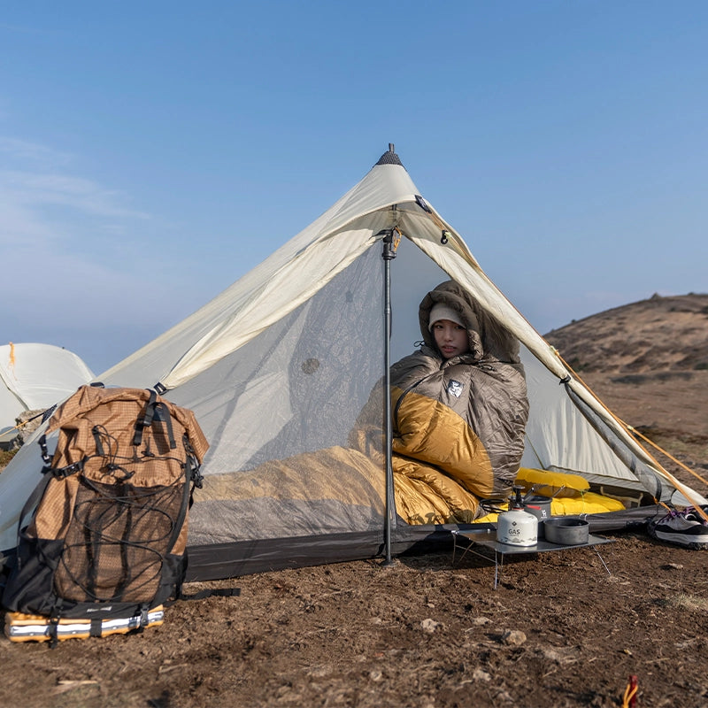 A girl wearing a sleeping bag sitting in a tent outdoors with a down yellow ultralight camping pillow next to her