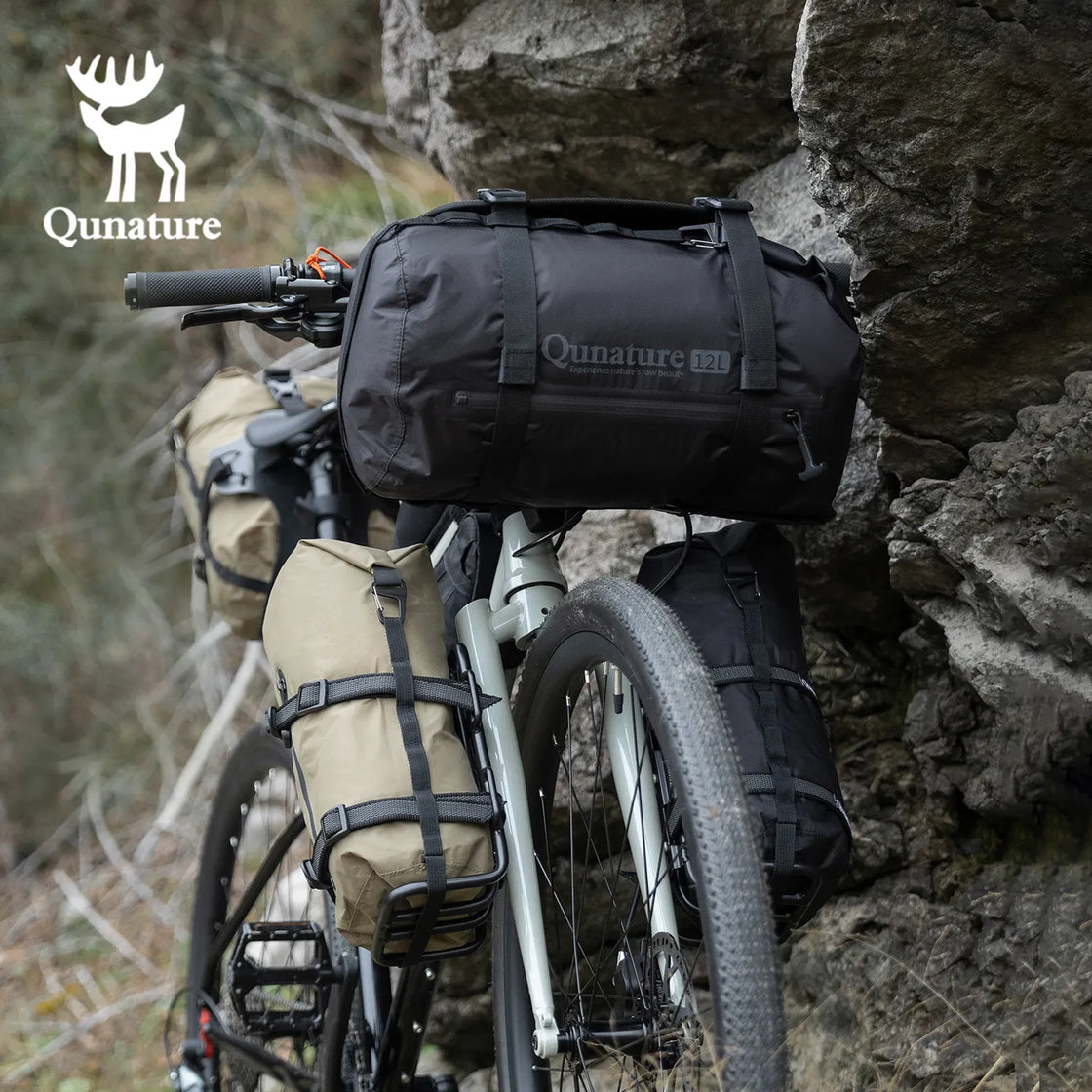 A black 12L large capacity waterproof compression bag is strapped to a gray bicycle next to the mountain