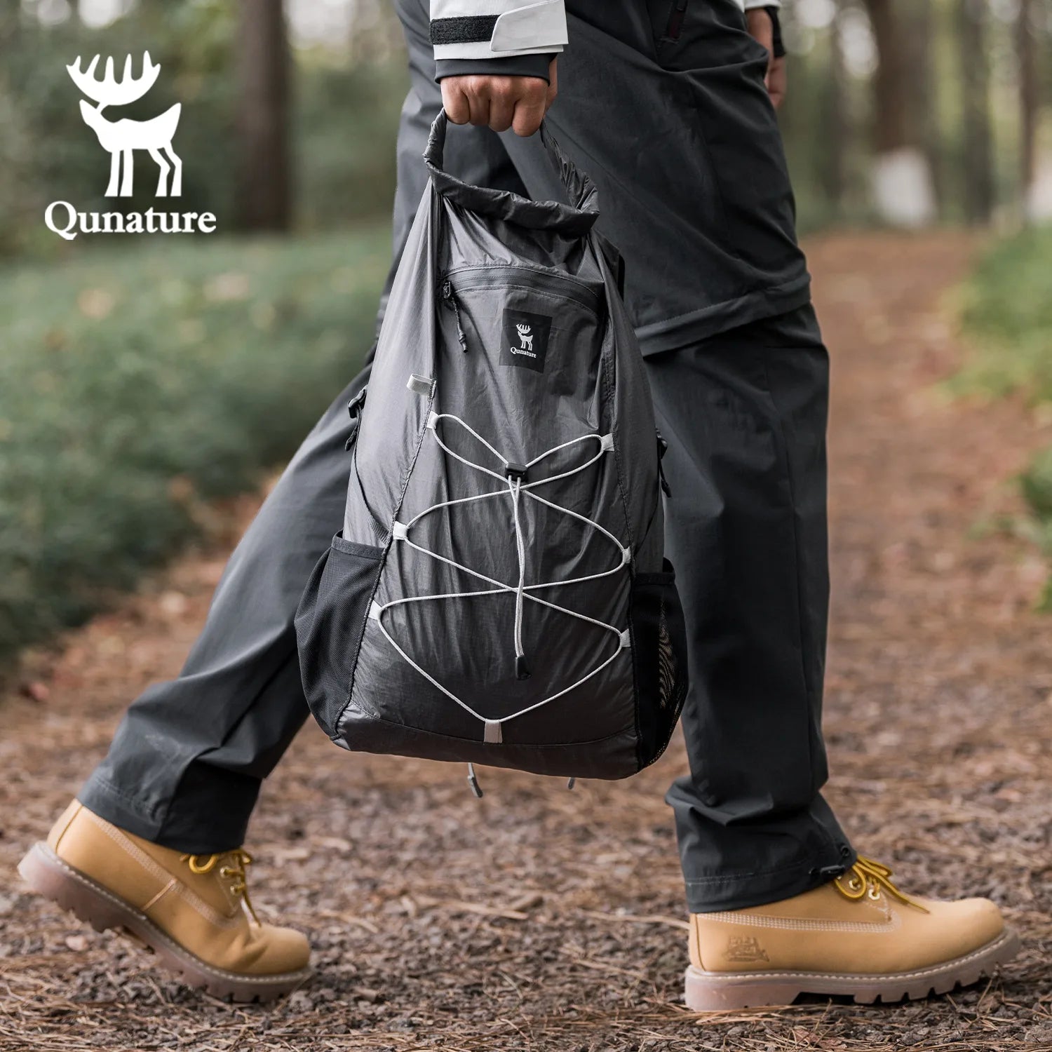 Person wearing dark pants and tan boots holding a black bag in a forest setting.